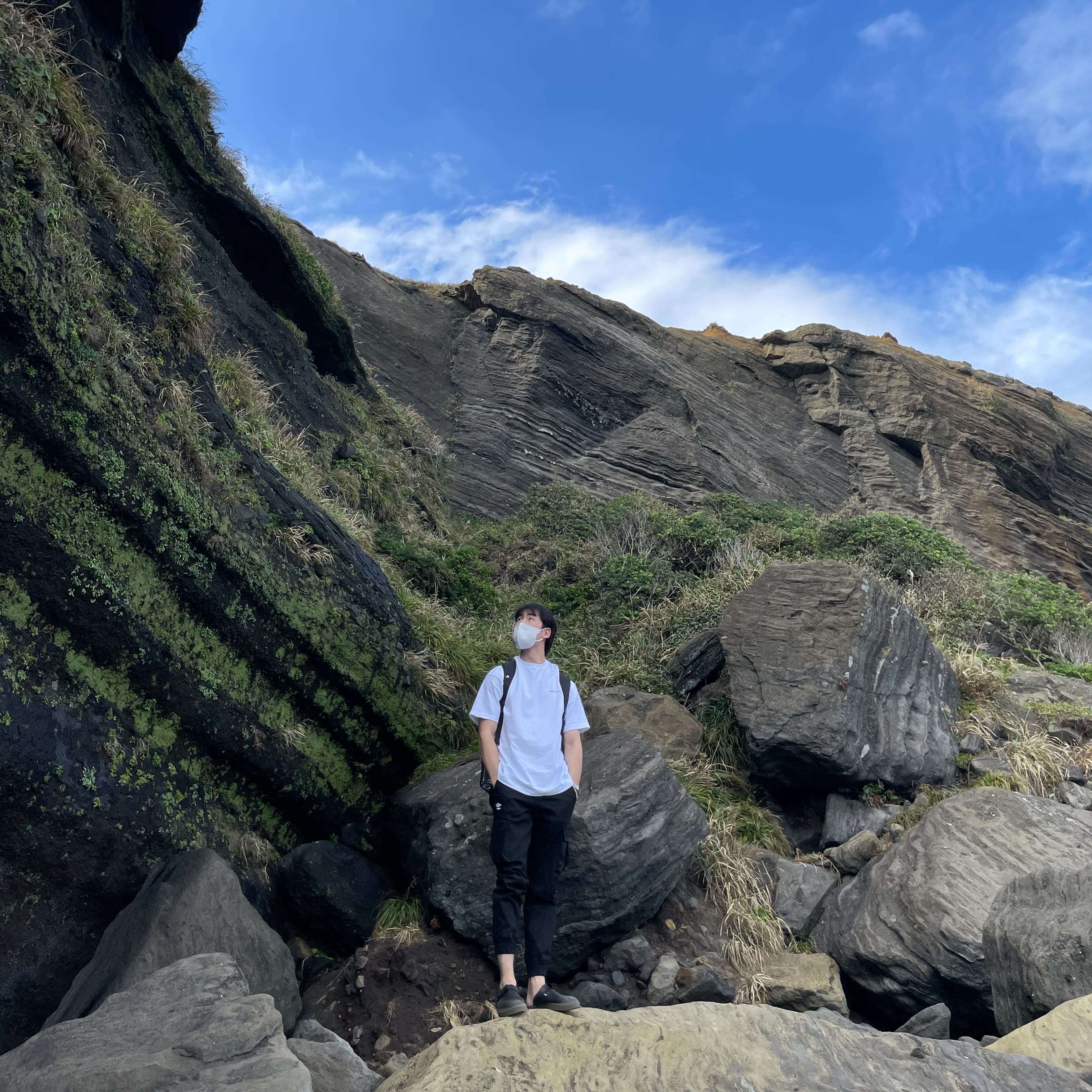 Image of In Tae standing in in front of a rocky wall and a blue sky above.